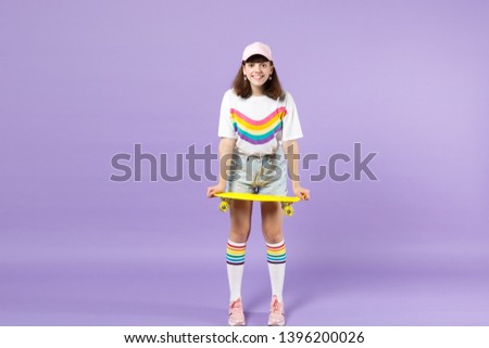 Portrait of joyful teen girl in vivid clothes standing, holding yellow skateboard isolated on violet pastel wall background in studio. People sincere emotions, lifestyle concept. Mock up copy space