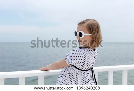 Small (8 years old) pretty cheerful girl in a white dress with black polka dots is standing on a wooden white pier. Selective focus.