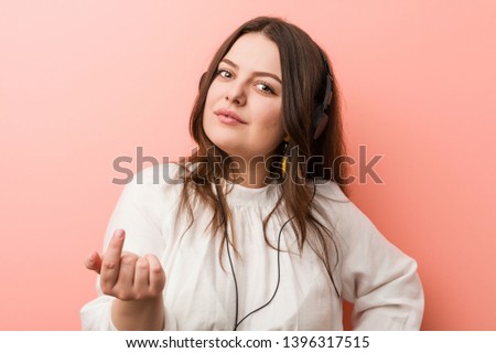 Young plus size curvy woman listening music with headphones pointing with finger at you as if inviting come closer.