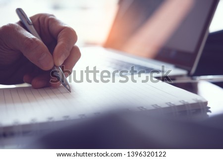 Business man hand write his work plan on paper notebook with a pen while working in modern office. Man student taking note while learning online course via laptop computer, close up, elearning concept