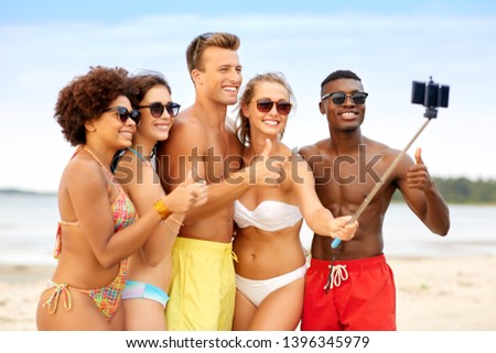 friendship, summer holidays and people concept - group of happy friends taking picture by smartphone on selfie stick on beach and showing thumbs up