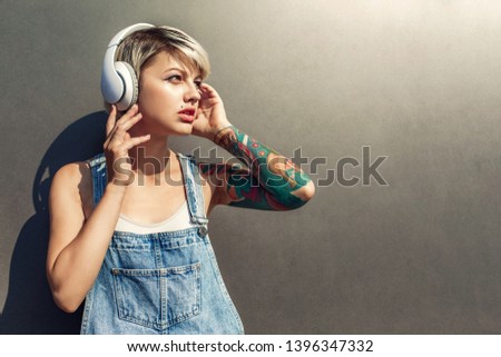 Young alternative girl wearing headphones standing isolated on grey wall on the city street listening to music looking aside pensive close-up