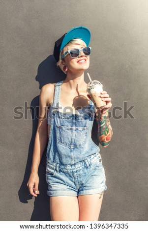 Young alternative girl wearing cap and sunglasses standing isolated on grey wall on the city street holding cup drinking iced coffee looking up smiling cheerful
