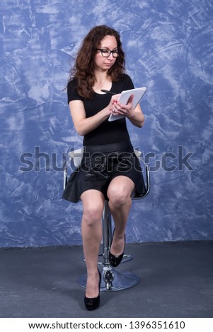 A young woman in black clothes is sitting on a chair on a blue background. Business woman with pen and notepad.