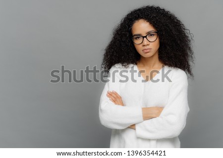 Self confident dark skinned ethnic female keeps hands crossed over chest, looks seriously at camera, wears optical spectacles and white sweater, stands against grey background with empty space
