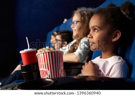 Beautiful african girl with funny hairstyle watching excited movie in cinema. Wonderful little child sitting with friends, eating popcorn and smiling. Concept of entertainment and enjoyment. Royalty-Free Stock Photo #1396360940
