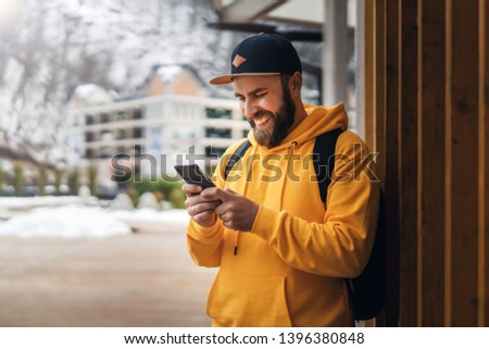 Front view. Bearded male tourist in yellow hoodie and cap with black backpack stands outdoors and using smartphone. Hipster man checks e-mail on smartphone, browses social networks. Lifestyle.
