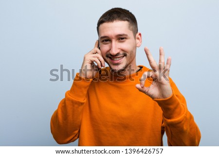Young caucasian man holding a phone cheerful and confident showing ok gesture.