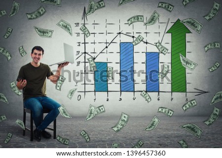 Young man sitting on chair holding laptop in one hand and mobile phone in another, celebrate business success, showing a positive increasing graph of virtual sales and money falling as a dollar rain.