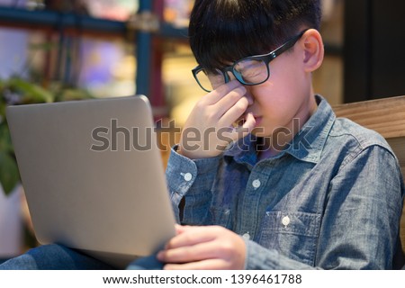 Smart looking tween / preteen Asian boy wears blue light blocking glasses from LED computer, squeezes his nose as his eyes strained, stressed from long screen exposure.  Computer Vision Syndrome (CVS) Royalty-Free Stock Photo #1396461788