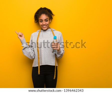 Young fitness black woman pointing to the side with finger.Holding a water bottle.