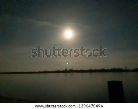 A blurred photo of the moon above the river and forest on the opposite bank.