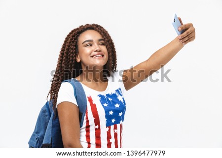 Portrait of cheery african american woman wearing backpack smiling and taking selfie photo on cellphone while standing isolated against white wall