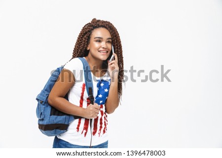 Portrait of cute african american woman wearing backpack smiling and talking on cellphone while standing isolated against white wall