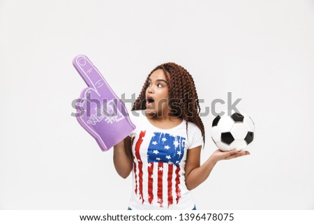 Portrait of energetic african american woman holding number one fan glove and soccer ball while standing isolated against white wall