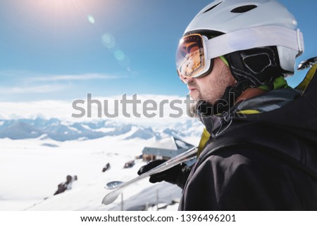 Close-up athlete skier in helmet and ski mask against the snow-capped mountains of a ski resort with a reflection of the Caucasian mountains in the mask