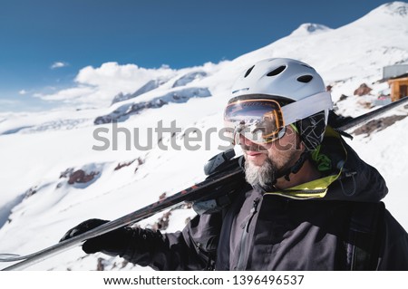 Close-up Portrait bearded male skier aged against background of mountains. An adult man wearing ski googles mask and helmet skis on his shoulder looks mountains. Ski resort concept