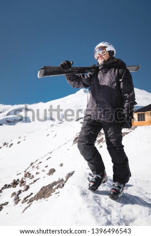 Growth Portrait bearded male skier aged against background of snow-capped Caucasus mountains. An adult man wearing ski googles mask and helmet skis on his shoulder looks mountains. Ski resort concept