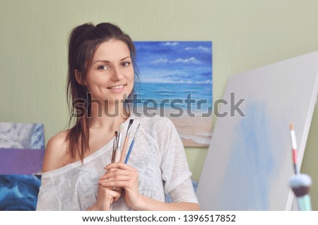 A beautiful painter holds many different brushes to paint while standing near the canvas at home against the background of a painting of the sea.