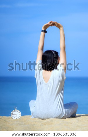 Young asian women with headphones, listening to music at the beach. White alarm clock on the sand. Blue sea and sky background.Concept for relaxation, travel vacation and holidays .Objects in sand.