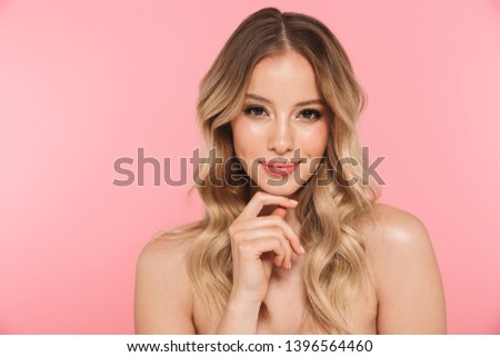 Portrait of a smiling beautiful young girl with blonde curly hair standing isolated over pink background, posing, touching face