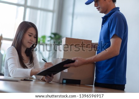 Young Asian woman signing on paper before receiving product package from delivery service company staff.