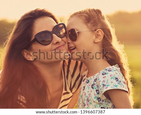 Happy fashion kid girl whispering her mother the secret in trendy sunglasses in profile view and looking on nature background. Closeup portrait of happiness. Toned portrait