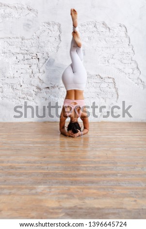 Concentration and preparation. Fit, unidentified young woman in sports suit makes stand on  head with her legs crossed - yoga exercise in an empty gym with white wall background. Copy space Royalty-Free Stock Photo #1396645724