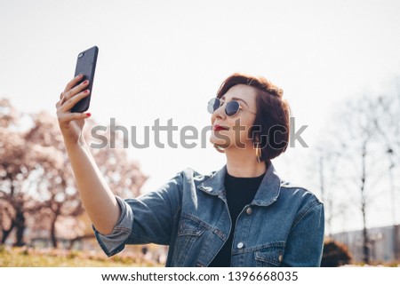 Candid portrait of stylish young woman taking a selfie with mobile phone, posing for social media in a park outdoors.