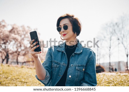 Candid portrait of stylish young woman taking a selfie with mobile phone, posing for social media in a park outdoors. 