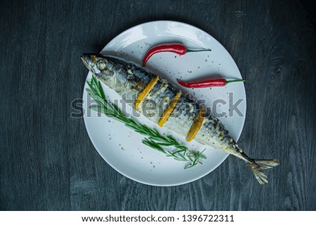 Baked mackerel with lemon on a white plate. Decorated with fresh rosemary, chili pepper. View from above. Dark wooden background.