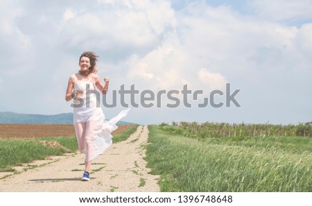 Girl in a vintage dress. Running along the picturesque spring hills at sunset. A long train of white skirt fluttered on the run. Photo of a happy brunette girl.  - Image 