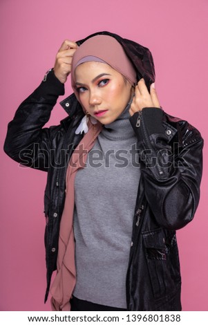 Fashionable female model in black pants, long sleeves leather hoodie jacket and hijab isolated on pink background. Stylish Muslim female hijab fashion lifestyle portraiture concept.