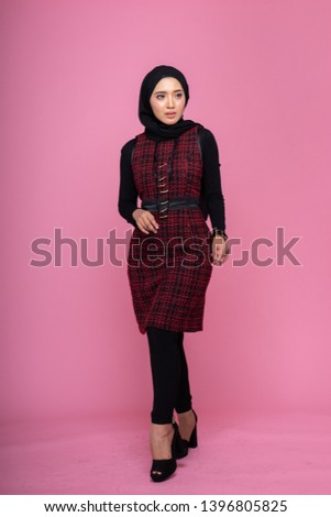 Fashionable female model in black jeans, long sleeves dress with hijab isolated on pink background. Stylish Muslim female hijab fashion lifestyle portraiture concept.