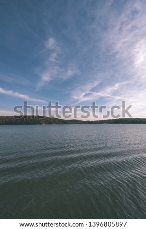 blue sky white clouds over calm body of water in summer with green foliage, trees and grass on the shores - vintage retro look