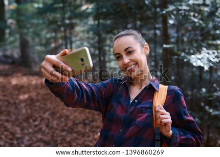 Beautiful happy stylish traveling woman taking photo on the camera phone with forest background. Portrait of young female hiker walking on the trail in the woods.