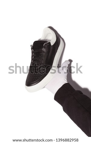 Male hand holding black leather sneakers over white background. Man presents black and white footwear isolated on white. Male hand in white glove holding  black shoes