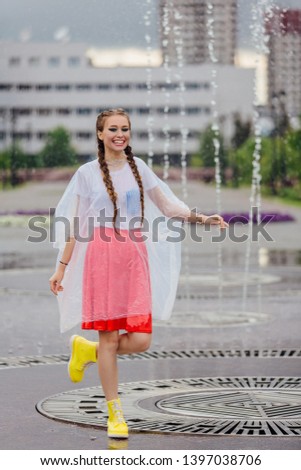 Young wet pretty girl with two braids in yellow boots and with transparent umbrella stands inside of fountain. Rainy day in city.