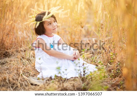 Little Asian girl sitting on ground in grass field, Kid in white dress and flower crown sitting alone in the field with warm sunlight in the evening