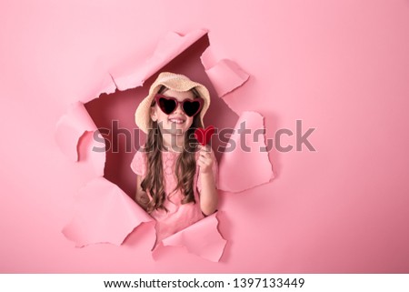 funny little girl looks out from the hole in a beach hat and glasses in the shape of a heart, holding a heart on a stick, on a colored background, a place for text, Studio shooting