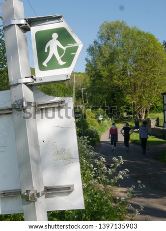Footpath sign showing white man person symbol on green arrow background beside tarmac path with people on it amongst hedges in rural gloucester england 