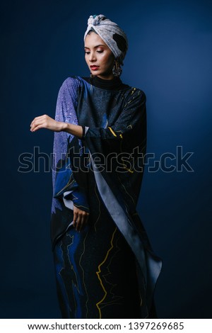Studio portrait of a young, elegant, tall and slim Middle Eastern Muslim woman wearing traditional clothing for Eid. She is in a flowing dark blue dress and matching turban (hijab head scarf).  Royalty-Free Stock Photo #1397269685