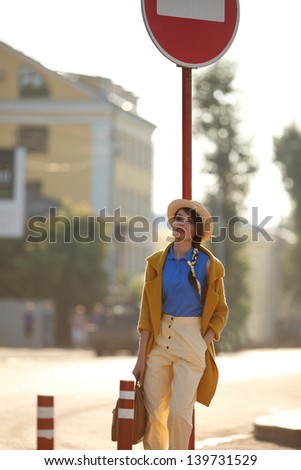 Young happy funny (vintage) dressed woman with retro suitcase stands on the street near red small columns and road sign.  Picture ideal for illustating woman magazines.