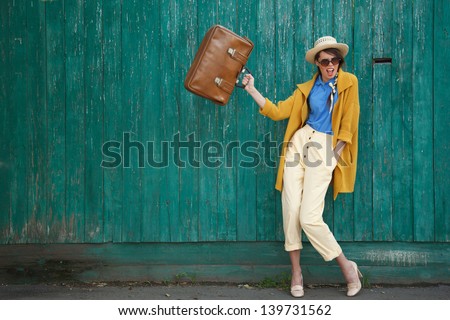Young happy funny (vintage) dressed woman waves retro suitcase. Old green fence on the background.  Picture ideal for illustating woman magazines.