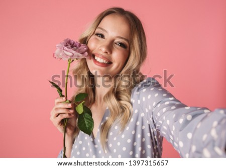 Beautiful young blonde woman wearing summer dress standing isolated over pink background, taking a selfie, holding a rose