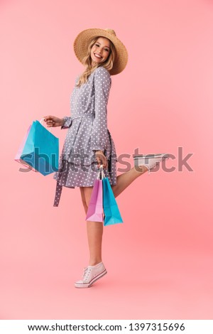 Full length portrait of a beautiful young blonde woman wearing summer straw hat standing isolated over pink background, holding shopping bags