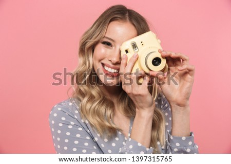 Portrait of a beautiful young blonde woman wearing summer dress standing isolated over pink background, using instant photo camera