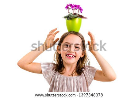 Girl carrying flower pot on head, having fun smiling. Little girl in dress holding green flower pot with viola flowers on her head having fun, goofing around. Studio shot isolated on white background