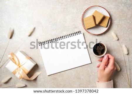 Modern minimal home workspace desk with blank paper notebook, human hand holding coffee cup, waffles, dry flowers on concrete stone background. Flat lay style, top view fashion lifestyle blog hero