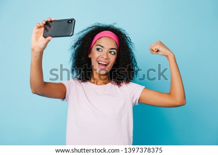 Laughing black girl showing muscle while posing on blue background. Studio shot of young african woman taking selfie.